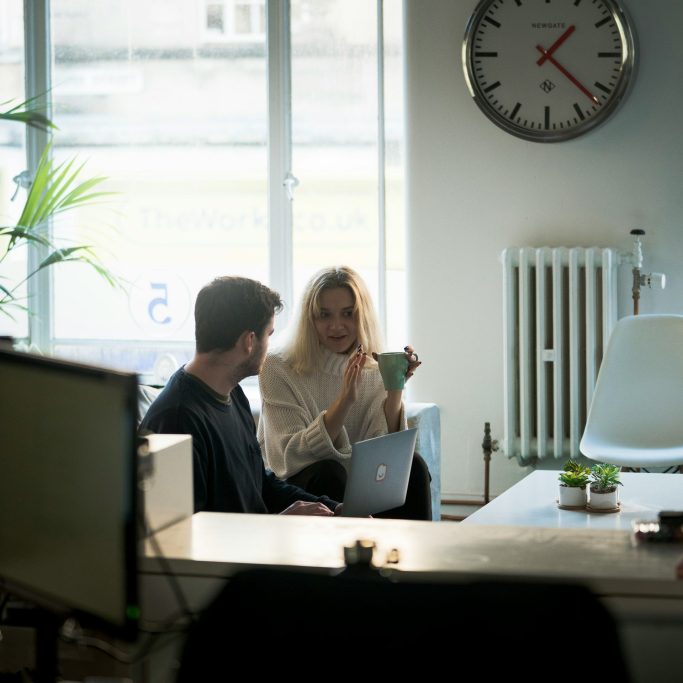 Två personer sitter i ett kontor, en pratar medan den andra arbetar på en laptop.